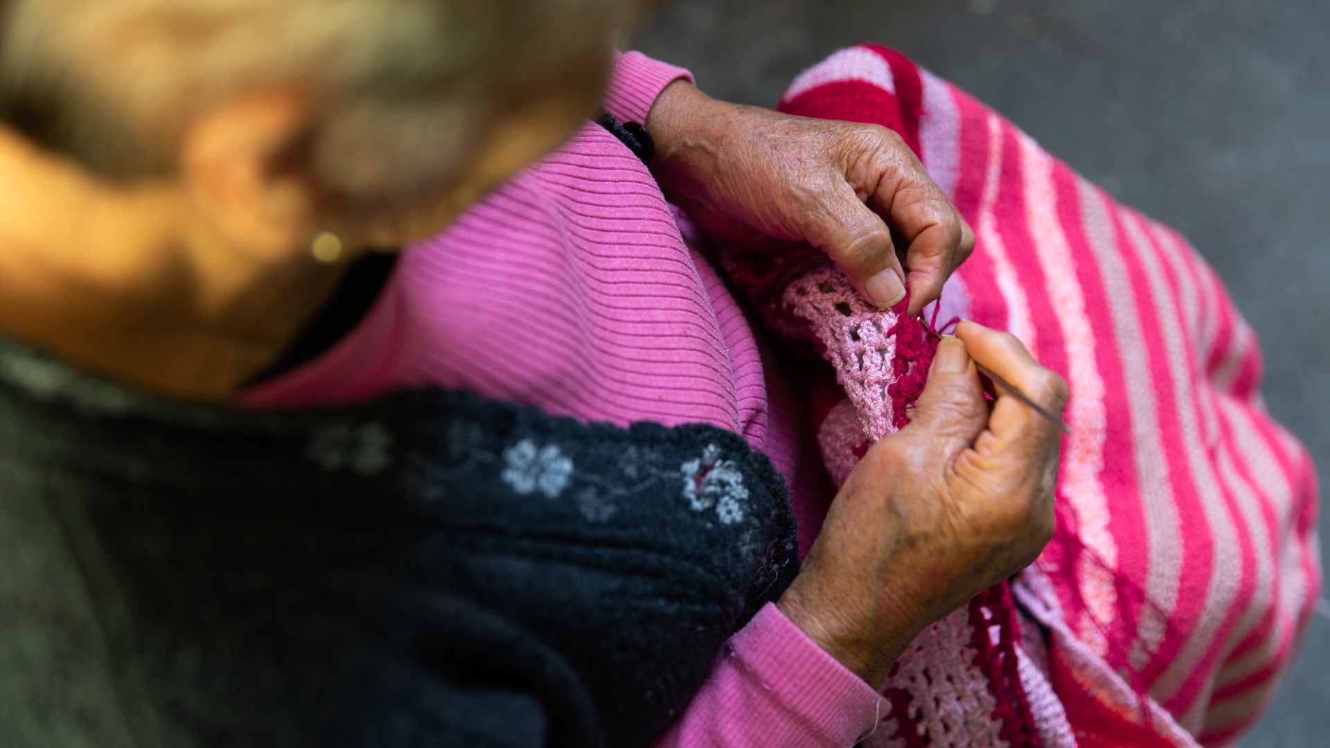 En gammal person sitter och virkar något randigt i utomhusmiljö, varmt solsken.