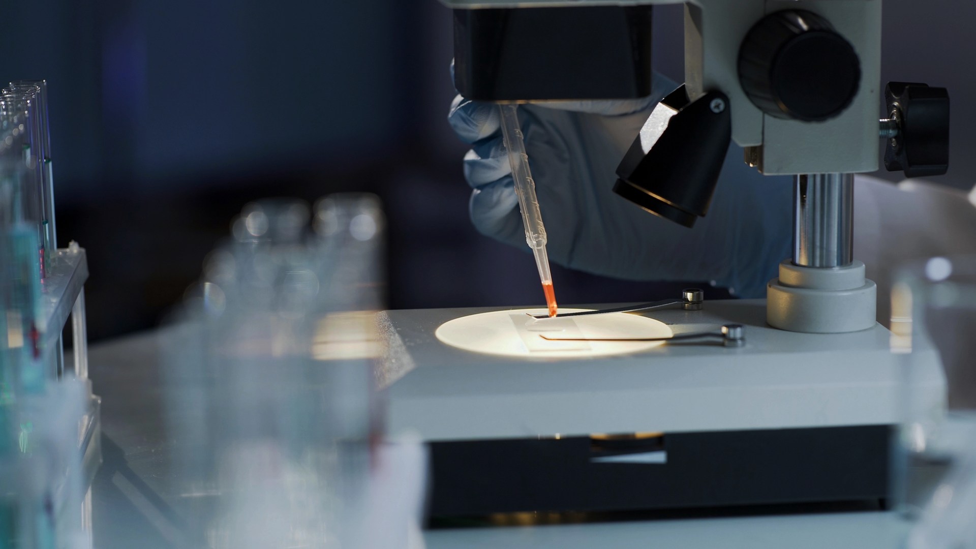 Labbmiljö med en hand med pipett som droppar en röd vätska på ett glas i ett mikroskop.