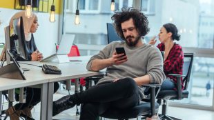 Ung man med mörkt skägg och mörkt lockigt hår sitter på skrivbordsstol och tittar på sin mobiltelefon. Två kvinnor sitter på varsin sida om kontorsbord och pratar, i bakgrunden.