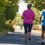 Två äldre personer joggar i lummig omgivning.