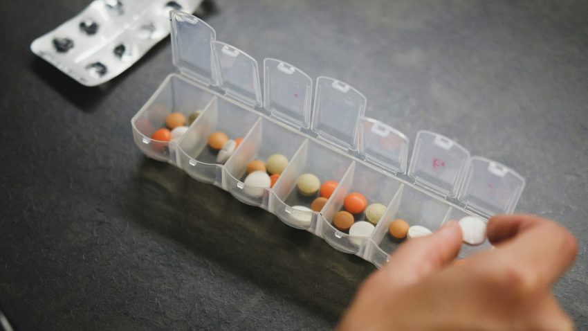 En medicindosa med många fack där det ligger piller i olika färger. En hand håller i ett piller.