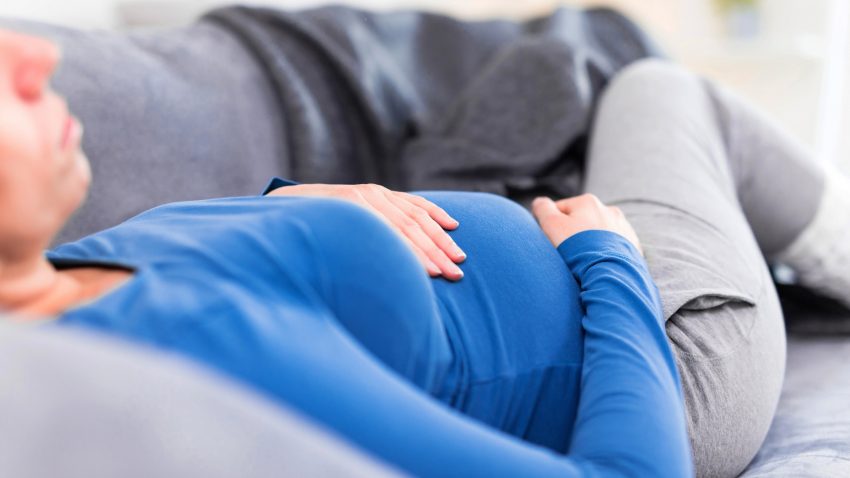 Trött gravid kvinna med blå tröja vilar i en soffa.