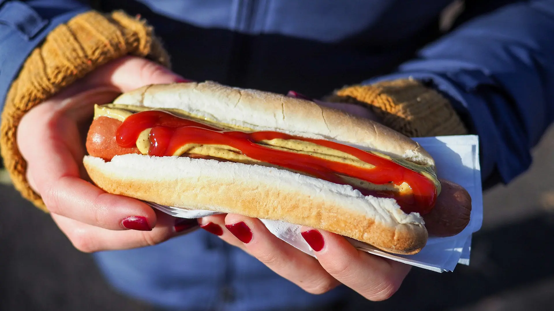 Två händer håller i varmkorv med bröd och ketchup och senap.
